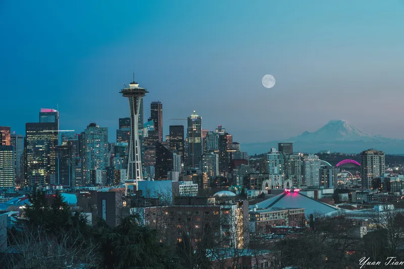 Full moon rising over the Seattle skyline at blue hour, city lights on and Mount Rainier in the distance