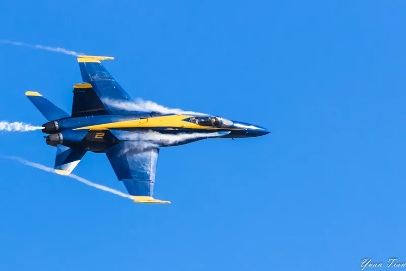 US Navy Blue Angels F/A-18 in a high-speed pass, vapor streaming over its wings against a deep blue sky
